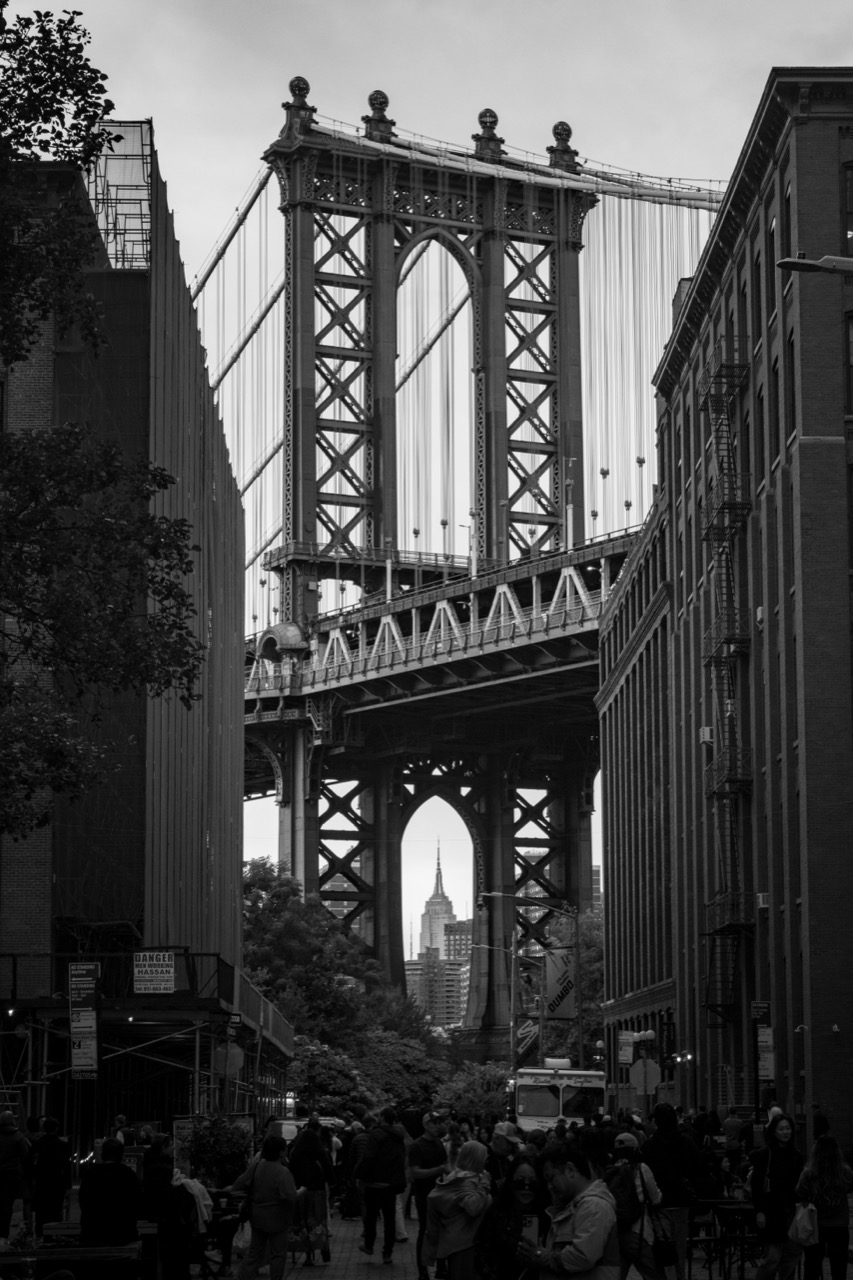 Black and white view of the Manhattan Bridge framed by buildings in DUMBO, with the Empire State Building beyond